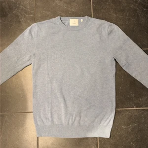 Ljusblå tröja från Bläck, storlek S - En stilren ljusblå tröja från Bläck i storlek S. Tröjan har rund halsringning, långa ärmar och ribbade muddar vid ärmslut och nederkant. Perfekt för lager-på-lager och enkel att matcha med jeans eller chinos.