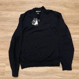 Svart half zip tröja från BOSS - Snygg svart långärmad tröja från BOSS med stilren half zip-dragkedja vid halsen. Tröjan är i mjuk bomull och har diskret BOSS-logga på bröstet. Perfekt för dig som gillar en clean och klassisk look.
