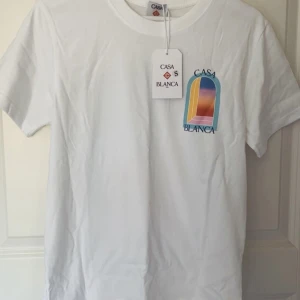 Vit t-shirt från Casablanca med tryck - Vit t-shirt från Casablanca med färgglatt grafiskt tryck fram och bak. Klassisk passform, rund hals och korta ärmar. Materialet är mjuk bomull och trycket har blå, gul, orange och rosa toner. Perfekt för dig som gillar stilrena men ändå unika plagg.