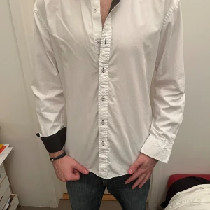 Vit skjorta med svarta detaljer - Snygg vit skjorta med svarta kontrastdetaljer på insidan av kragen och manschetterna. Skjortan har klassisk krage, långa ärmar och knappar framtill samt på manschetterna. Materialet känns som bomull och har en stilren look med coola svarta sömmar vid knapphålen.