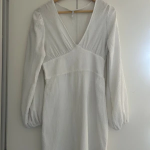 Vit kortklänning med v-ringning från ONLY - Snygg vit kortklänning från ONLY med v-ringad hals och långa puffärmar. Klänningen har en lätt rynkad struktur i tyget och smock i ryggen för extra komfort. Perfekt för dig som gillar stilrena och ljusa plagg med feminin touch.