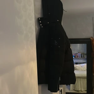 Svart pufferjacka från Moose Knuckles - Säljer en svart pufferjacka från Moose Knuckles med ikonisk logga i metall på ärmen. Jackan har stor huva med tryckknappar, dragkedja framtill och på ärmslut samt flera fickor. Riktigt varm och skön. 