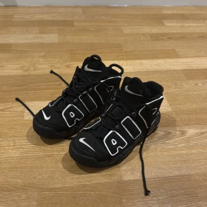 Nike Air More Uptempo Svarta Sneakers - Svarta Nike Air More Uptempo sneakers med feta vita AIR-bokstäver på sidorna och klassiska Swoosh-detaljer. Skorna har snörning, hög siluett och en chunky sula. Tillverkade i syntet och skinn för en sportig vibe. Perfekta för dig som vill sticka ut med en ikonisk streetstyle.