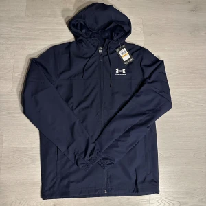 Marinblå vindjacka från Under Armour - Snygg marinblå vindjacka från Under Armour med huva och justerbar snörning. Jackan har dragkedja framtill, elastiska muddar och vit logga på bröstet. Perfekt för dig som gillar sportig stil och vill ha en lätt jacka med clean look.