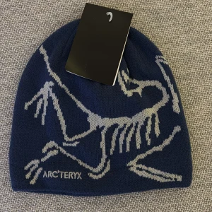 Blå mössa från Arc'teryx med fossilmotiv - Snygg marinblå mössa från Arc'teryx med ett stort fossilmönster i ljusgrått. Mjuk och skön stickad kvalitet som passar perfekt när det är kallt ute. Loggan är broderad framtill och designen är riktigt clean och modern.