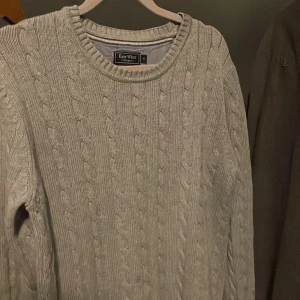 Ljusgrå kabelstickad tröja East West - Kabelstickad tröja från East West i ljusgrå färg. Rund halsringning och ribbade muddar vid ärmslut och nederkant. Stickad struktur med klassiskt mönster, perfekt för dig som gillar enkel och stilren look.