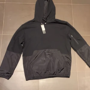 Svart vindjacka från J.Lindeberg - Svart vindjacka från J.Lindeberg med huva och snörning. Jackan har en mix av material med slät polyester på nedre delen och ärmarna, samt en stor ficka framtill. Långärmad och stilren design med dragkedja på ena ärmen.