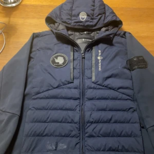 Mörkblå Sail Racing pufferjacka - Snygg mörkblå pufferjacka från Sail Racing med huva och coola patchar på bröstet och ärmen. Jackan har dragkedja framtill och quiltad design. Perfekt för dig som gillar sportig stil och vill ha något som sticker ut. finns några hål som har blivit fixade