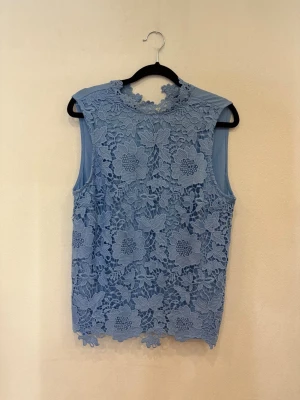 Blå ärmlös blus med spetsdetaljer - Supersöt ljusblå ärmlös blus med blommig spets på ryggen och runt halsen. Framsidan är slät medan baksidan har ett vackert spetsmönster som ger en romantisk vibe. Perfekt för dig som gillar detaljer och vill sticka ut lite extra.