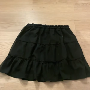 Svart volangkjol från Sister, S - Svart kortkjol från Sister i storlek S med flera lager volanger och elastisk midja. Kjolen är gjord i ett lätt, syntetiskt material som ger ett snyggt fall och extra volym. Perfekt för dig som gillar en trendig och feminin look. 