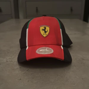 Ferrari x Puma röd keps - Snygg Ferrari keps från Puma i rött och svart med detaljer i grönt och vitt. Framsidan har Ferrari-loggan och skärmen har Puma-märket. Kepsen har klassisk form och är tillverkad i polyester, perfekt för dig som gillar motorsport och vill sticka ut. Aldrig använd äkta köpt i Italien 