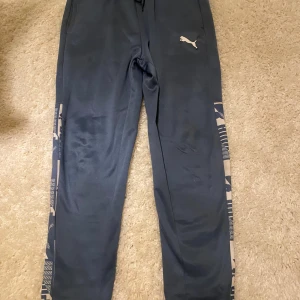 Grå mjukisbyxor från Puma M - Snygga grå mjukisbyxor från Puma i storlek M. Byxorna har beige detaljer längs sidorna med grafiskt mönster och Puma-logga framtill. Tillverkade i mjuk polyester med elastisk midja och snörning för skön passform.Qr coden leder till Pumas hemsida