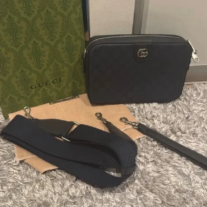 Blå axelväska från Gucci i skinn - Snygg svart axelväska från Gucci i skinn med diskret GG-mönster och silverfärgade detaljer. Väskan har avtagbar och justerbar axelrem samt dragkedja upptill. Perfekt för dig som vill ha en stilren och lyxig accessoar.