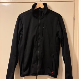 Svart vindjacka från Sail Racing - Svart vindjacka från Sail Racing med hög krage och hel dragkedja framtill. Jackan har en diskret bröstficka med dragkedja och en rund Sail Racing-patch på ärmen. Materialet är mjukt och syntetiskt, perfekt för blåsiga dagar. Kommer utan luva 