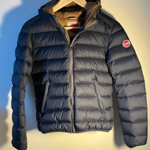 Mörkblå Colmar dunjacka med huva - Snygg mörkblå dunjacka från Colmar med huva och dragkedja framtill. Jackan har ett quiltat mönster och två sidofickor. Insidan är beige och har en stor Colmar-logga. Perfekt för kalla dagar och har en clean, sportig vibe.