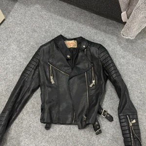 Svart skinnjacka med dragkedjor - Säljer en svart skinnjacka i storlek 36 från Vogather med coola detaljer som quiltade partier på axlar och ärmar, flera silvriga dragkedjor och bälten i midjan. Jackan har en snygg asymmetrisk front och långärmade ärmar med zip. Perfekt för dig som gillar edgy stil. Jackan är använd och har några slitage endast om man kollar nogrannt 