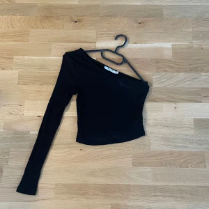 Svart oneshoulder topp från NA-KD - Säljer en svart ribbad oneshoulder topp från NA-KD i storlek XS. Toppen har lång ärm på ena sidan och är croppad i modellen, vilket ger en snygg och modern look. Perfekt för dig som gillar trendiga och stilrena plagg.