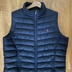 Mörkblå dunväst från Polo Ralph Lauren stl XS - Snygg mörkblå dunväst från Polo Ralph Lauren med horisontella quiltade paneler och röd broderad logga på bröstet. Västen har hög krage och dragkedja framtill, perfekt för lager-på-lager.  Storleken är XS och passar 160cm 