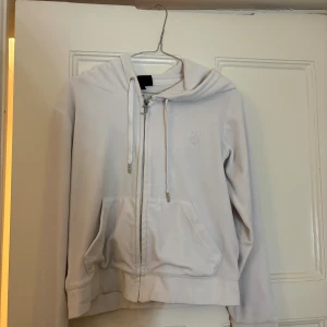 Vit hoodie från Juicy Couture - Vit hoodie från Juicy Couture med dragkedja och snyggt J-hänge. Tröjan har huva med snörning, två fickor fram och broderad logga på bröstet. Mjuk bomullskänsla och långärmad, perfekt för chill dagar.