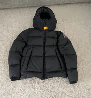 Svart dunjacka Parajumpers Endurance - Säljer en svart Parajumpers Endurance dunjacka med huva och gul detalj vid halsen. Jackan har en quiltad design, dragkedja framtill och patch på ärmen. Insidan är grå med praktisk ficka. Perfekt för kalla dagar och riktigt snygg stil. Ny pris 7000kr.