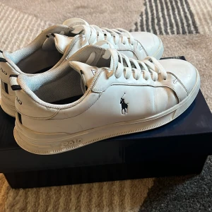 Vita sneakers från Polo Ralph Lauren - Fräscha vita sneakers från Polo Ralph Lauren med klassisk låg profil och rund tå. Skorna har snörning, svart Polo-logga på sidan och svarta detaljer på hälen. Tillverkade i skinn med vit platt sula och diskret branding på sulan och hälen.