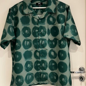Grön mönstrad skjorta från Stüssy - Snygg kortärmad skjorta från Stüssy i grönt med coolt prickigt mönster. Skjortan har klassisk krage, bröstficka och knappar framtill. Materialet känns lätt och luftigt, perfekt för varmare dagar. Passformen är normal och designen sticker ut med sitt unika print.