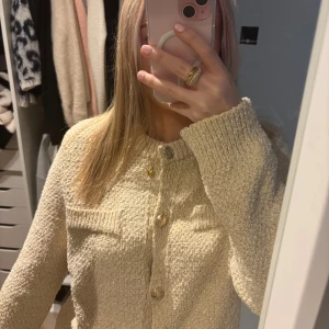 Beige stickad kofta med gulddetaljer - Superfin beige stickad kofta med glittriga gulddetaljer i materialet och snygga guldfärgade knappar framtill. Koftan har rund hals, långa ärmar och två dekorativa fickor på bröstet. Perfekt att slänga över en topp för en chill look.