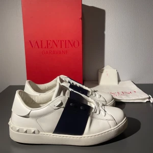 Valentino Garavani Open sneakers vit/blå - Snygga Valentino Garavani Open sneakers i vitt skinn med bred mörkblå rem över vristen och coola nitar på hälen. Klassisk rund tå, vita snören och platt sula. Kommer med originalkartong och dustbag. Perfekta för dig som gillar lyxiga detaljer och stilren design.