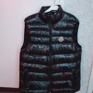 Svart dunväst från Moncler - Snygg svart dunväst från Moncler med glansig finish och horisontella quiltade paneler. Västen har hög krage, dragkedja framtill och två fickor med dragkedja. Moncler-logga på bröstet ger en exklusiv touch. Perfekt för lager-på-lager under kyliga dagar.