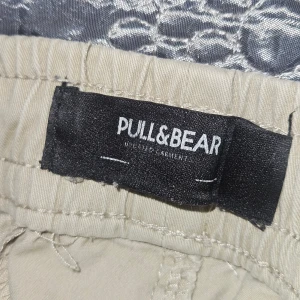 Beige cargopants från Pull&Bear - Säljer ett par beige cargopants från Pull&Bear med elastisk midja och snörning. Byxorna har flera praktiska fickor på sidorna och baktill, samt resår vid bensluten för en chill och avslappnad look. Perfekt för dig som gillar streetwear och funktionella detaljer.