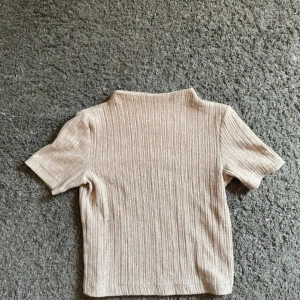 Beige ribbad topp från Zara - Säljer en beige ribbad topp från Zara med korta ärmar och hög hals. Toppen har en slim passform och är tillverkad i ett mjukt, stickat material som känns skönt mot huden. Perfekt att styla med jeans eller kjol för en clean look.