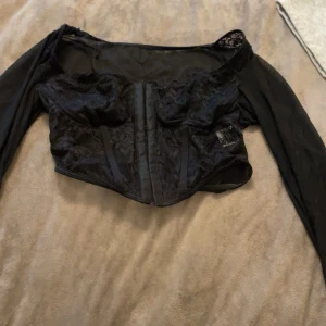 Svart spetskorsett med långa ärmar - Säljer en svart korsett-topp med långa ärmar och snygga spetsdetaljer. Toppen har croppad passform och stängning framtill med hakar. Spetsen ger en cool och elegant vibe, perfekt för att lyfta din outfit. Materialet är mjukt och följsamt. Pris kan diskuteras och köparen står för frakt