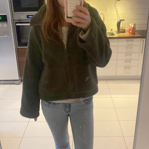 Mörkgrön teddyjacka med dragkedja - Säljer en mysig mörkgrön teddyjacka med dragkedja framtill och stor krage. Jackan har en loose passform och är perfekt för kyliga dagar. Materialet är fluffigt och ger en skön känsla. Passar dig som gillar avslappnad stil och vill ha något varmt och trendigt. Knappt använd. 