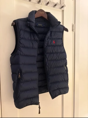 Marinblå dunväst från Polo Ralph Lauren - Snygg marinblå dunväst från Polo Ralph Lauren med röd logga på bröstet. Västen har quiltad design, hög krage och två praktiska fickor med dragkedja. Perfekt att slänga över en hoodie för extra värme under höst och vinter.