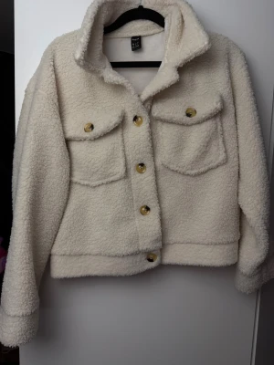 Vit teddyjacka från Shein - Mysig teddyjacka i vit färg från shein, med krage och stora bröstfickor. Jackan har guldfärgade knappar framtill och är tillverkad i fluffigt fleece-material. Modellen är croppad och har lång ärm, perfekt för att ge en chill vibe till din outfit.