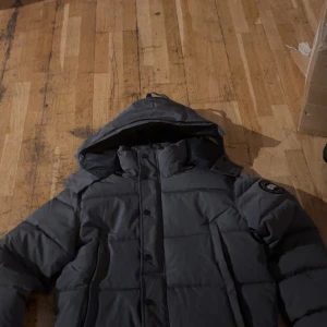 Mörkgrå dunjacka Canada Goose - Säljer en mörkgrå dunjacka från Canada Goose med huva och klassisk patch på ärmen. Jackan har quiltad design, dragkedja och knappar framtill. Perfekt för kalla dagar och har en riktigt schysst streetstil. Fodrad insida och snygg passform.