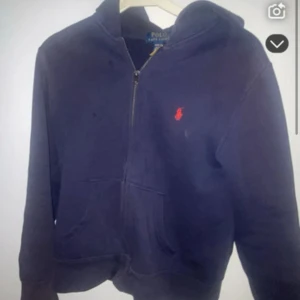 Mörkblå hoodie från Polo Ralph Lauren - Snygg mörkblå hoodie från Polo Ralph Lauren med röd broderad logga på bröstet. Tröjan har huva, dragkedja framtill och två fickor. Perfekt för chill dagar och enkel att matcha med jeans eller joggers.
