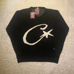 Corteiz stickad tröja - Svart stickad Corteiz tröja med stor vit C och stjärna på framsidan. Rund hals och ribbade muddar vid ärmslut och nederkant. Långärmad och mjuk, perfekt för att hålla värmen. Snygg detalj med liten broderad logga vid nederkanten.