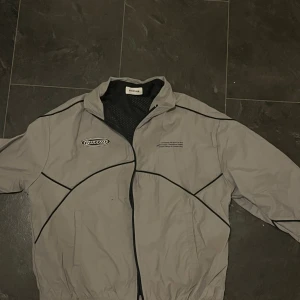 Grå vindjacka från YOUTH LUV - Säljer en grå vindjacka från YOUTH LUV med svarta detaljer och coola sömmar. Jackan har dragkedja framtill, elastisk nederkant och meshfoder på insidan. Märkeslogga på bröstet och textprint ger jackan en sportig vibe. Perfekt för dig som gillar streetwear.