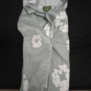 Grå hoodie med vita blommor Denim Tears - Grå hoodie från Denim Tears med vita blommor tryckta över hela tröjan. Tröjan har huva vilket gör den både chill och praktisk. Perfekt för dig som gillar streetwear med en twist. Materialet är mjuk bomull för skön komfort.