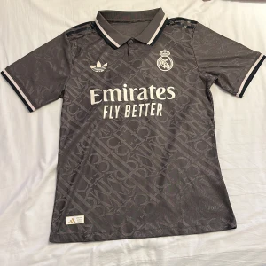 Grå Real Madrid Vini Jr. fotbollströja - Snygg grå Real Madrid fotbollströja från Adidas med Vini Jr. och nummer 7 på ryggen. Tröjan har korta ärmar, krage och subtilt mönster i tyget. Tryck med Emirates Fly Better på bröstet och klubbens emblem. Perfekt för dig som älskar fotboll och streetstyle.