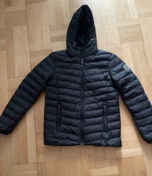 Svart pufferjacka från Vinson Polo Club - Svart pufferjacka från Vinson Polo Club med huva och dragkedja framtill. Jackan har ett quiltat mönster och två sidofickor. Märkets logga syns på ärmen. Perfekt för kyliga dagar och har en stilren look.