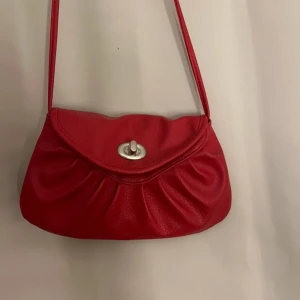 Röd axelväska i skinn med silverdetalj - Snygg röd axelväska i skinn med veckad framsida och silverfärgat vridlås. Väskan har ett långt axelband och ett fack med dragkedja inuti. #vintage #bag