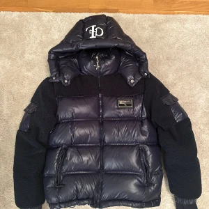 Svart pufferjacka från Chriztian Lane - Snygg svart pufferjacka från Chriztian Lane med stor huva och logga framtill. Jackan har en cool mix av glansigt syntetmaterial och teddyfleece på axlar och ärmar. Flera fickor med dragkedja och patchdetaljer på ärmarna. Riktigt snygg och cool