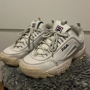 Fila Disruptor Low vita chunky sneakers - Fila Disruptor Low sneakers i vitt med chunky plattformssula och snörning. Ikonisk Fila-logga på sidan och plösen, robust design med grov yttersula för extra attityd. Tillverkade i syntetmaterial och skinn, perfekt för dig som gillar streetstyle.