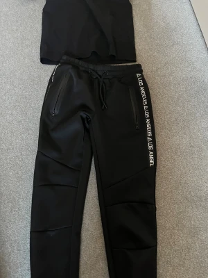 Svarta mjukisbyxor Chapter Young och en tröja  - Svarta mjukisbyxor från Chapter Young med coolt Los Angeles-tryck längs benet. Byxorna har snörning i midjan, dragkedjor på fickorna och är tillverkade i ett mjukt syntetmaterial. Perfekt för chill eller streetwear-look.