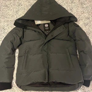 Mörkgrå dunjacka från Canada Goose - Säljer en mörkgrå dunjacka från Canada Goose med huva och diskret logga på ärmen. Jackan har dragkedja och knappar framtill, två stora fickor och ribbade muddar vid ärmsluten. Perfekt för kalla dagar och riktigt snygg passform.