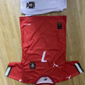 Portugal röd fotbollströja Puma - Röd Portugal fotbollströja från Puma med nummer 7 och namn på ryggen. Tröjan har små vita mönster, gröna och vita detaljer på ärmsluten samt Portugals landslagsemblem på bröstet. Tillverkad i lätt och ventilerande material.