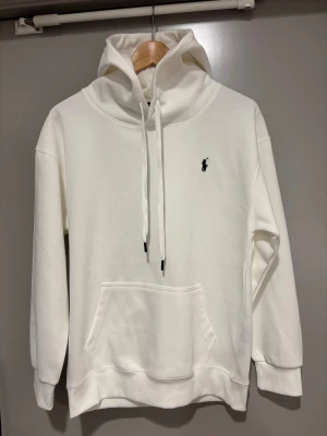 Vit hoodie från Polo Ralph Lauren - Vit hoodie från Polo Ralph Lauren med klassisk svart logobrodyr på bröstet. Tröjan har huva med snörning, stor magficka och ribbade muddar. Mjuk bomullskvalitet och relaxed fit, perfekt för chill dagar. Snygg och enkel design som alltid funkar. Aldrig använd, bara hängt i garderoben