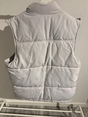 Ljusgrå dunväst med hög krage - Snygg ljusgrå dunväst från Zara Man med quiltad design och två stora fickor framtill. Västen har en hög krage och stängs med dragkedja och tryckknappar. Perfekt för lager-på-lager och chill stil under kyliga dagar.